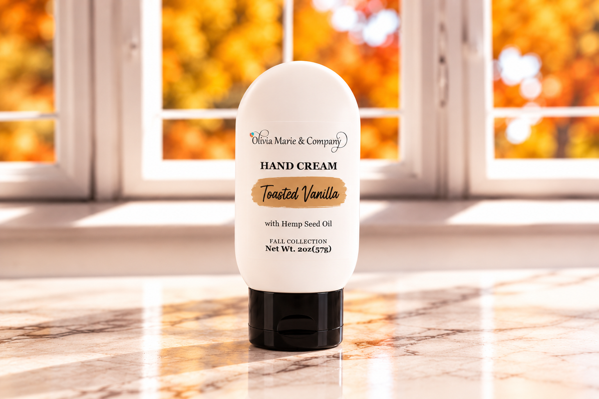 A white squeeze tube of hand cream with a black cap stands on a marble surface in front of a window with warm autumn foliage outside. The label reads 'Olivia Marie & Company HAND CREAM' and 'Toasted Vanilla'.