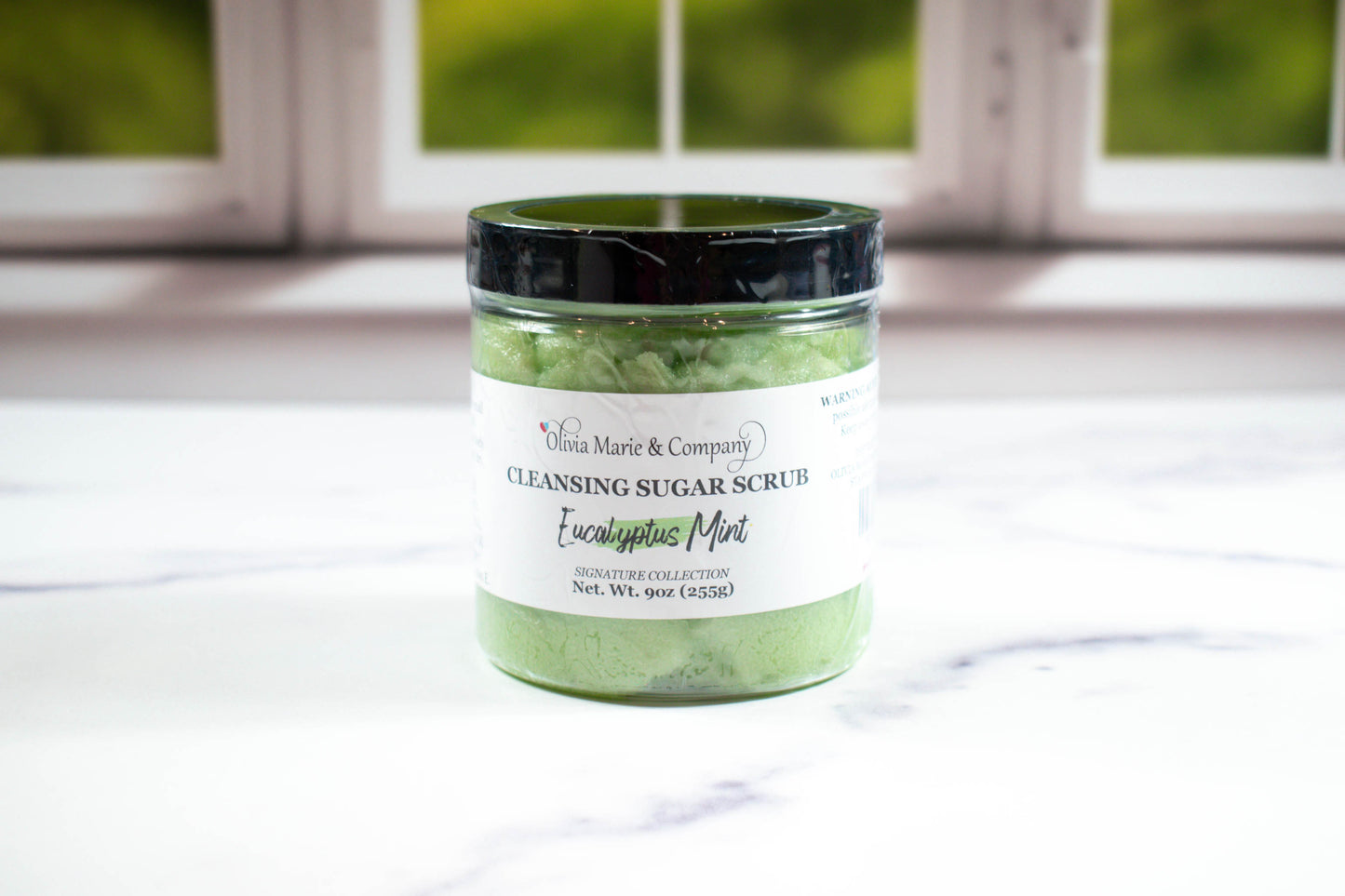 A jar of Eucalyptus Mint Cleansing Sugar Scrub placed on a marble surface with a window in the background.