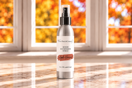 A silver room refreshier spray bottle with a black spray top and a white label reading 'Vivian Marie & Company' and 'Apple Harvest', standing on a marble countertop in front of a window with autumn foliage outside.