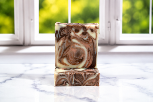 Two stacked rectangular bars of marbled artisan soap with brown, cream, and tan swirls, displayed on a white marble surface in front of a blurred window with green foliage outside.