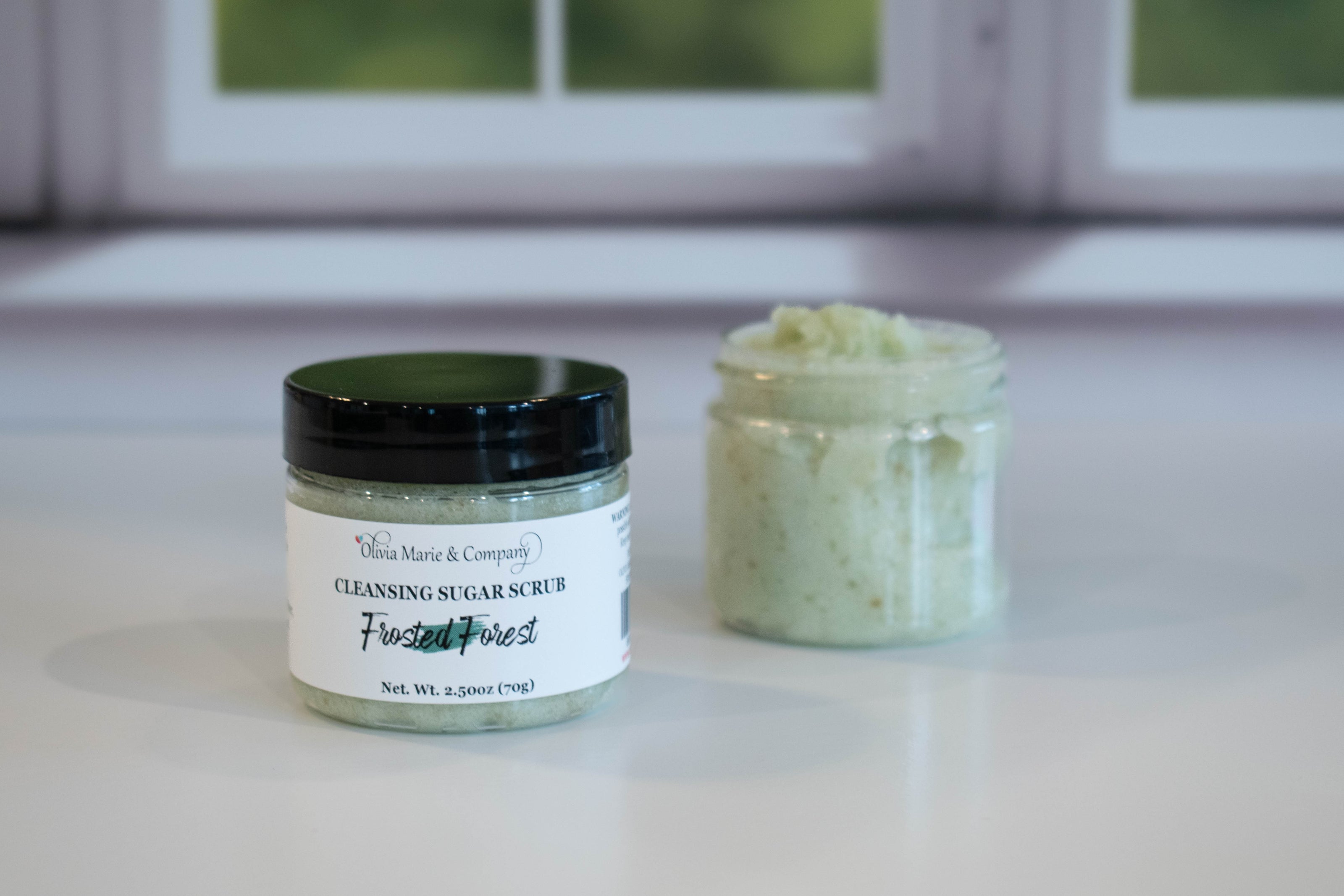 Two jars of cleansing sugar scrub on a white surface with a window in the background.