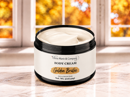 A jar of Olivia Marie & Company Golden Brulee Body Cream sits on a marble countertop with a blurred window showing autumn foliage in the background.