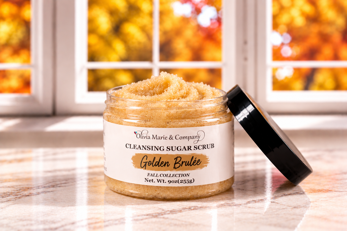 A jar of Olivia Marie & Company Cleansing Sugar Scrub in the Golden Brulee scent, shown open with its black lid leaning against it on a marble surface. The background features a window with a blurred autumn foliage view.